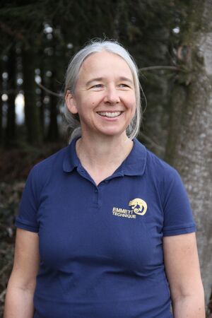 Eine lächelnde Person im blauen „Emmett Technique“ Poloshirt im Freien vor Bäumen.